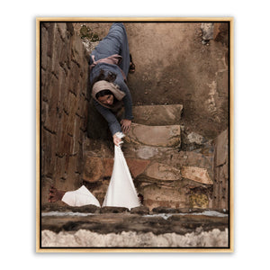 Aerial high-angle photograph looking down a stone staircase. A woman in a gray robe lies on the steps, arm extended, fingers reaching toward the visible hem of a white robe at the bottom edge of the frame.
